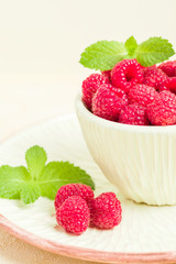 Ripe raspberries with green mint leaves in cup and saucer on pastel yellow background.