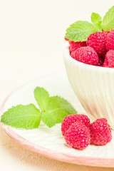 Ripe raspberries with green mint leaves in cup and saucer on pastel yellow background.