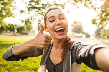 Photo of feminine fitness girl 20s in sportswear wearing bluetooth earpod taking selfie and showing...