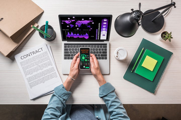 partial view of businessman holding smartphone with marketing analysis on screen at table with contract, stationery and laptop with infographic on screen
