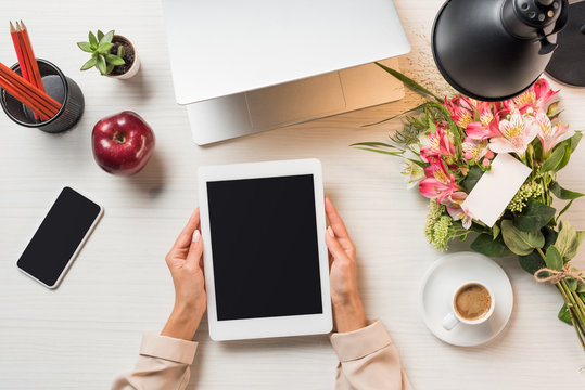partial view of female freelancer holding digital tablet with blank screen at table with coffee cup, gadgets and flowers with greeting card - Powered by Adobe