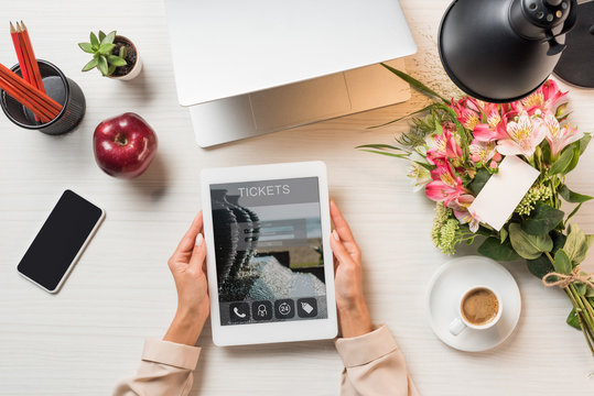 cropped image of female freelancer holding digital tablet with tickets on screen at table with coffee cup, gadgets and flowers with greeting card