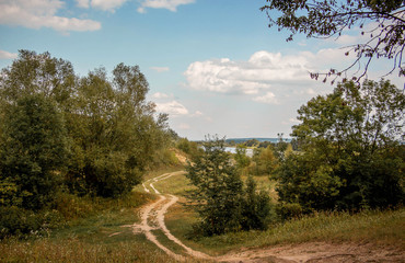 The winding dirt road between the trees. Autumn landscape with trees, road and blue sky with white clouds_