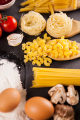 Fototapeta na wymiar Different types of uncooked raw macaroni and pasta next to egg in flour on dark background