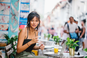 A young and trendy Chinese Asian millennial teenage girl wearing sunglasses and street clothes, sits and works on her laptop in a cafe. 