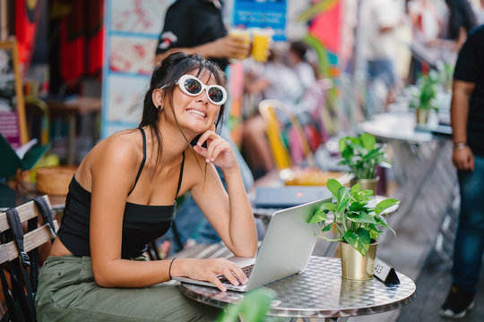 A young Chinese Asian girl is typing on her laptop while sitting at a cafe beside a crowded street on a sunny day. She is smiling while working.