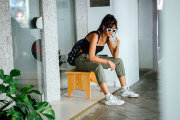 An image of a young, attractive, and athletic Asian teenage girl in a tank top and cargo pants. She is taking a break from exploring the city on a sunny day.