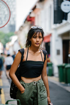 A Young Asian Millenial Is Walking Along The Street With Her Backpack In Tow. She Looks Serious And Nervous To Get To Her Destination.