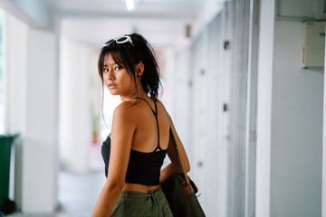 Portrait of a young, tanned, and athletic Asian teenage model girl in a tank top and cargo pants looking over her shoulder and smiling as she walks down a walkway in a street in the city.