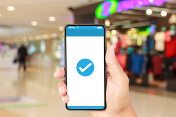Hand holding smartphone with a blue check mark icon on white screen over blurred in shopping mall background. Confirmed smartphone order success, Online payment concept