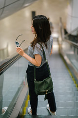 A young Japanese Asian woman is riding down the escalator. She is dressed in stylish casual clothing and smiling as she looks backwards at the camera. 