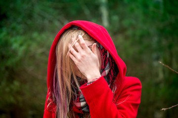 Girl with colorful hair in red autumn coat with hood is hiding and covering face with hand. Green...