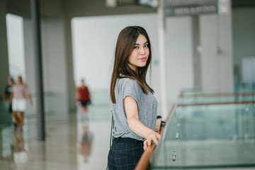 A young Japanese woman leans against the glass railing in a clean, futuristic mall. She looks amused by the view she is seeing.