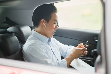 A young Japanese Asian man is talking on his smartphone using his bluetooth headset. He is riding at the back seat of a car he booked using a ride hailing app.