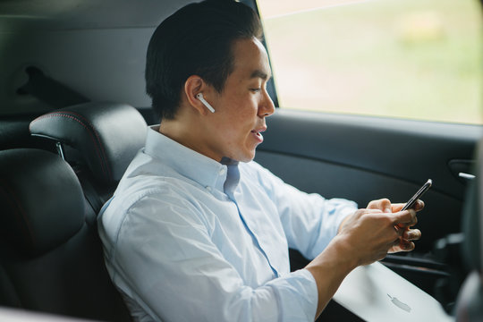 A Young Japanese Asian Man Is Talking On His Smartphone Using His Bluetooth Headset. He Is Riding At The Back Seat Of A Car He Booked Using A Ride Hailing App.