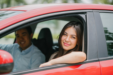 Portrait of a young Japanese Asian woman leaning out the window of a red car window and smiling happily. She is being driven to her destination in a ride she booked on a ride hailing app.