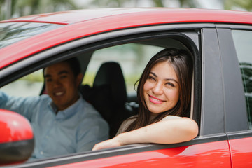 Portrait of a young Japanese Asian woman leaning out the window of a red car window and smiling happily. She is being driven to her destination in a ride she booked on a ride hailing app.