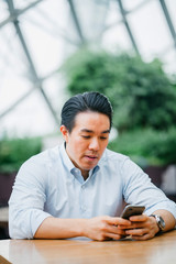 A portrait of a young Japanese man using his smartphone while seated outdoors on a bright day. He is wearing smart casual clothes and looks happy.