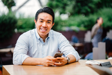 A portrait of a young Japanese man using his smartphone while seated outdoors on a bright day. He...