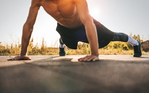 Cropped head image of young athletic muscular man doing push-ups on workout ground. Caucasian shirtless fitness male doing outdoor workout exercises on sunset light background. People, sport concept