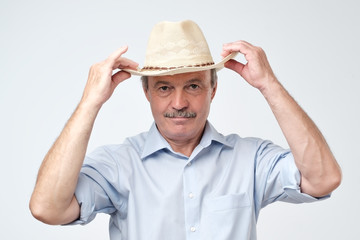 Cowboy style. Mature man adjusting his cowboy hat and looking at camera while standing against grey background