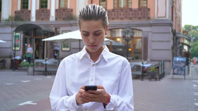 Young modern woman is standing on street and waiting for someone, typing message on smartphone, communication concept, urban concept