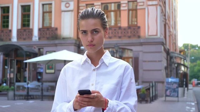 Young modern woman is standing on street and watching at camera, typing message on smartphone, communication concept, urban concept