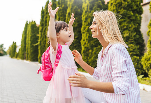 Horizontal image of happy little girl and mother on sidewalk next to the trees. Mom play together with toddler kid preschooler with backpack outdoor before kindergarten. Mother's day concept.