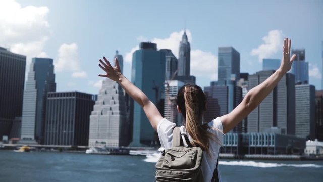 Back view of happy female tourist with backpack raising arms wide open at New York skyline view, looking back at camera.