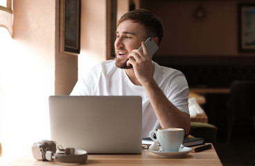 Young freelancer talking on mobile phone while working on laptop in cafe