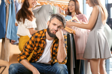 Annoyed young man waiting for his girlfriend and her friends while they choosing clothes in shop