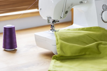 Close-up of a white sewing machine stitching a purple thread on a green fabric in a crafts room interior. Real photo.