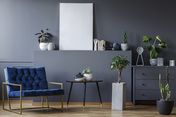 Real photo of a wide, blue armchair standing next to a table and a cupboard in a grey living room...