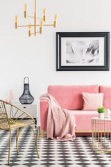 Poster above pink sofa with blanket in living room interior with gold chair and table. Real photo