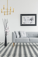 Plant next to grey couch in living room interior with checkered floor and poster. Real photo