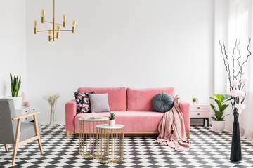 Patterned armchair near gold tables and pink sofa in white flat interior with plants. Real photo