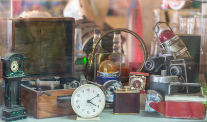 Assorted vintage items, clocks, cameras, flasks, sextant, lamps behind shop window.
