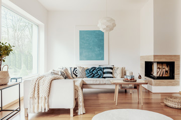 Turquoise blue knot pillow on a beige corner sofa and an abstract poster on a white wall in a...