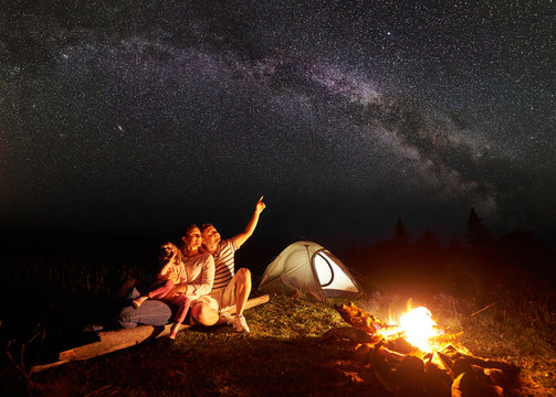 Family camping in mountains at night, sitting in front of illuminated tent and burning campfire. Woman holding in arms girl, man pointing at bright stars in dark sky. Tourism and traveling concept.