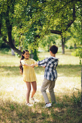 Obraz premium Sweet little couple spinning around holding hands. Beautiful young miss in summer yellow dress holding hands with young boy and spinning around in park.