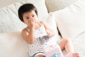 Sick little Asian girl wiping or cleaning nose with tissue sitting on sofa at home. Medicine and health care concept.