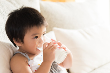 Happy little asian girl hand holding drinking milk glass, sitting on sofa at home. Medicine and health care concept.