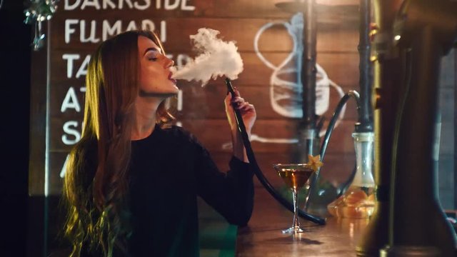 A girl drinks a cocktail and smoke the hookah at the bar counter in 4k resolution in slow motion