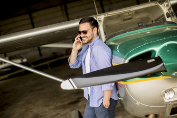 Handsome young pilot checking airplane in the hangar and using mobile phone