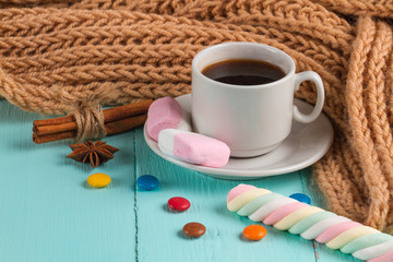 Obraz na płótnie Canvas A cup of coffee, a knitted, brown scarf, cinnamon, marshmallow, multicolored sweets and anisetree on a blue, wooden background. Drinks and sweets.
