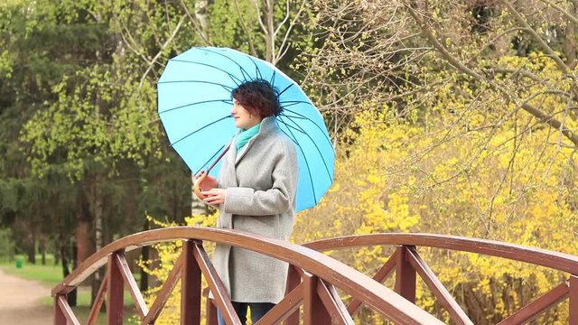 Pretty young red head woman with the blue umbrella on the bridge in spring park. Green trees and forsythia blossoms background.