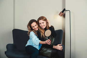 Two pretty young women sitting on sofa and looking to mirror in beauty salon. Two girl friends having fun together. Women fashion, makeup and hairstyle concept. Modern lifestyle.