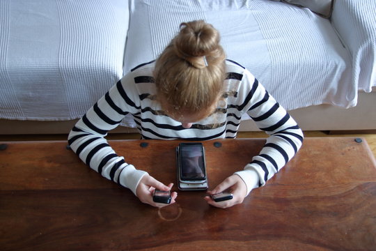 Młoda diewczyna, (dziewczynka) siedzi przy niskim drewnianym stole, na nim leży smartfone, dziewczynka w rękach trzyma jeszcze dwa telefony kom&oacute;rkowe, ma pochyloną głowę, patrzy na nie, blond włosy 