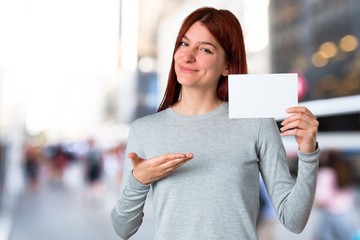 Young redhead girl holding an empty white placard for insert a concept on unfocused background