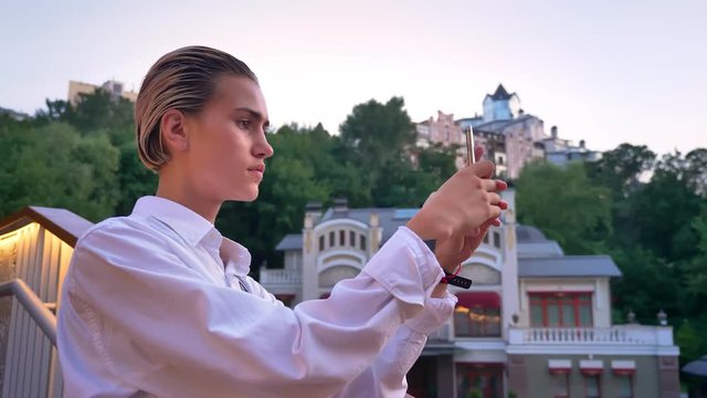 Young modern woman is standing on bridge, takes photo of sunset on smartphone, urban concept